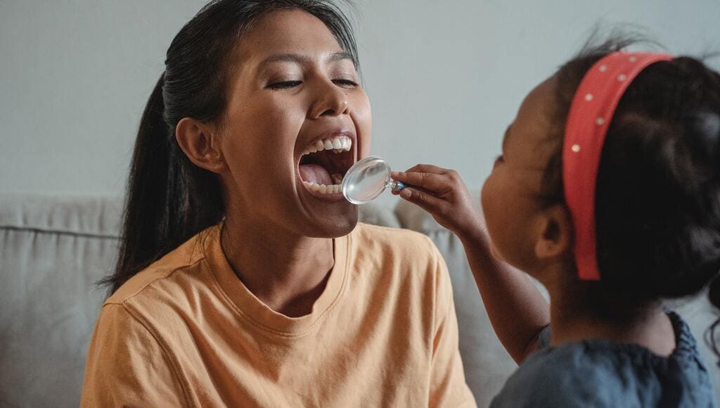 Criança examinando com lupa os dentes da mãe que mostra dentes perfeitos. Educação em saúde bucal começa na infância,