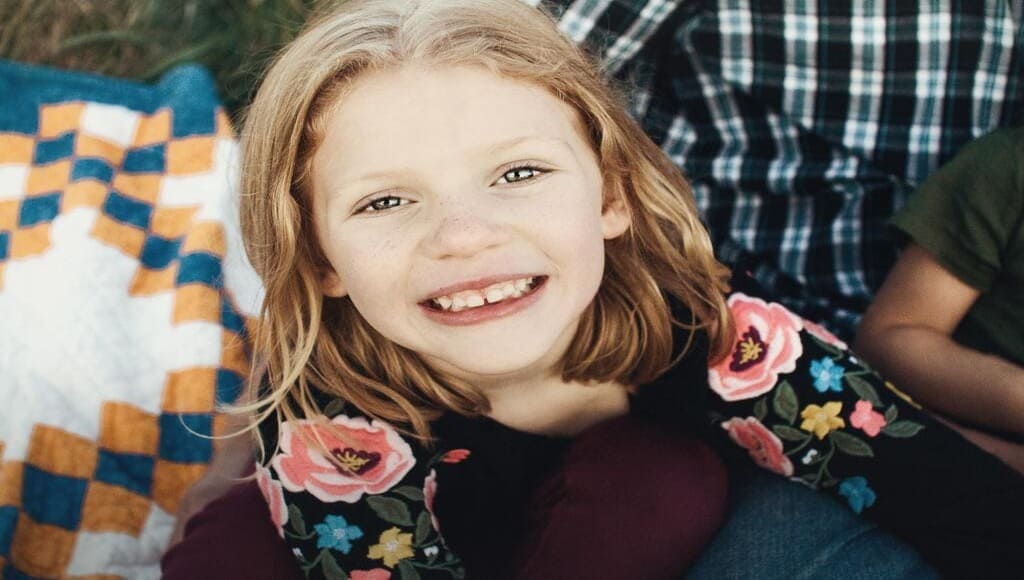 Menina ruiva de 9 anos, pele clara, roupas cor escura, jaqueta com flores de cor rosa, sorrindo e mostrando os dentes frontais muito afastados devido o freio labial anormal